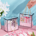 Two transparent black border gift bags with golden rope handles filled with gifts and pink shredded paper